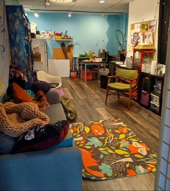 The Ratzon Space: a small room with chairs, a couch with blankets, and a rug in the foreground, and a table and shelves in the background. There is a sink in the back corner. The shelves are full and the back wall is painted blue.